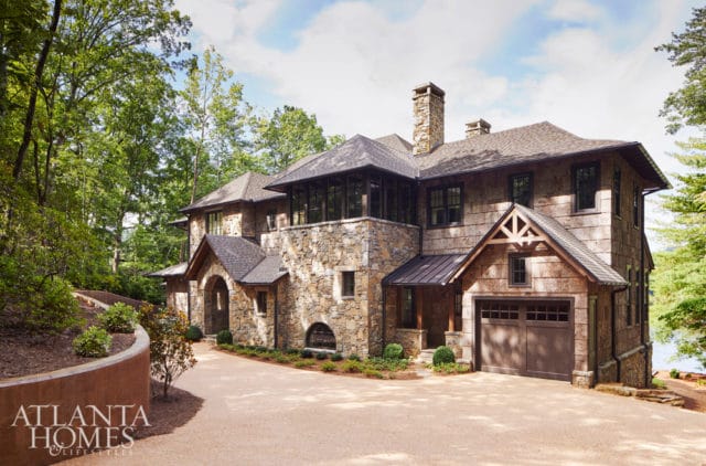Stone and wood shingle lake house with a multi-level roof, stone chimney, and attached garage, surrounded by lush green trees.