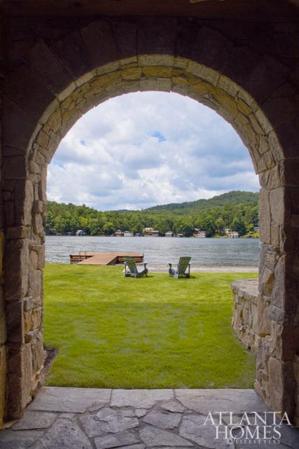 Stone archway overlooking a lake with a wooden dock, two Adirondack chairs on a green lawn, and tree-covered hills in the background.