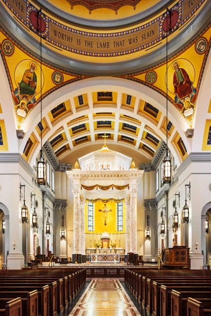 Interior of a cathedral with ornate gold and yellow ceiling murals, marble columns, pews, and a brightly lit altar featuring a crucifix.