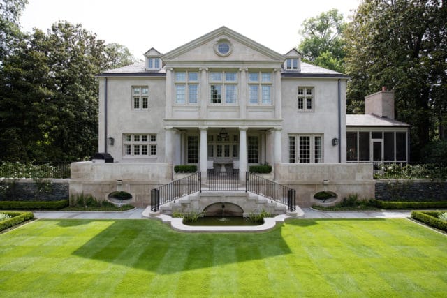 Neoclassical stone mansion with a grand entrance, symmetrical windows, and a screened porch, set within a manicured lawn and mature trees.