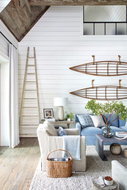 Living room with white shiplap walls, a light blue sofa, striped armchair, wooden ladder, and two rattan canoe-shaped wall decorations