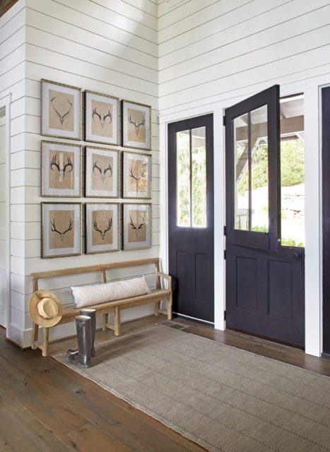 Entryway with shiplap walls, a gallery wall of framed deer antlers, a wooden bench with a bolster pillow, hat, and boots, and black double doors leading outside.