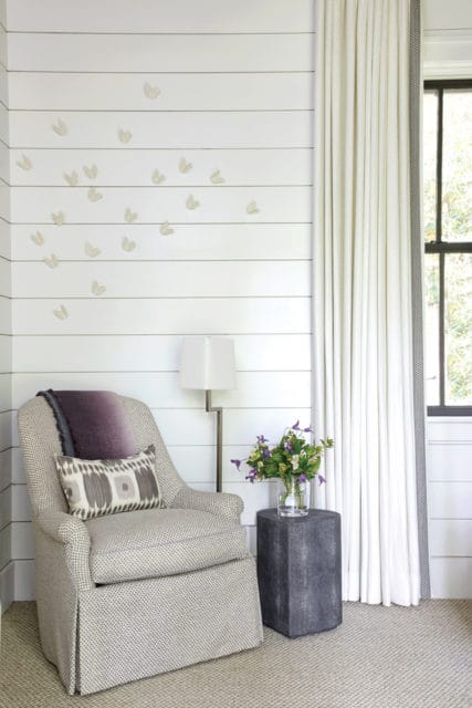 Armchair with patterned upholstery and purple throw pillow, next to a dark gray side table with a vase of flowers, against a white shiplap wall with butterfly decals.