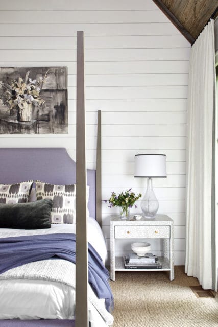 Bedroom with a four-poster bed, lavender upholstered headboard, shiplap walls, and a patterned nightstand with a glass lamp and floral arrangement.