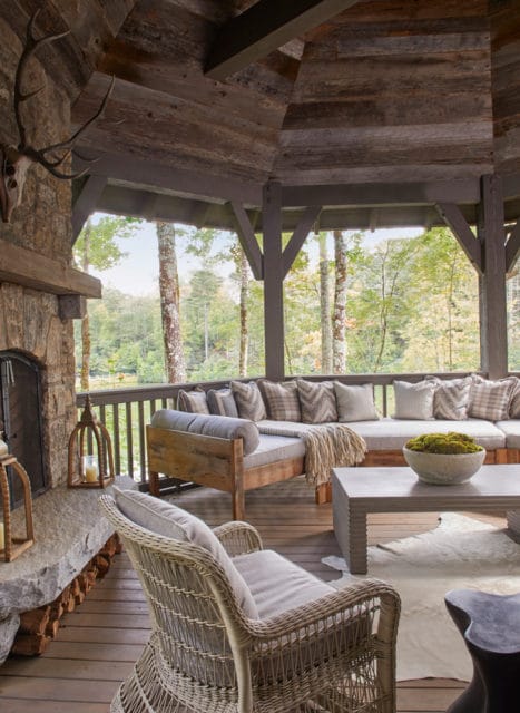 Covered porch with stone fireplace, rustic wood ceiling, and sectional sofa with plaid pillows. Wicker chair in foreground overlooks wooded landscape.