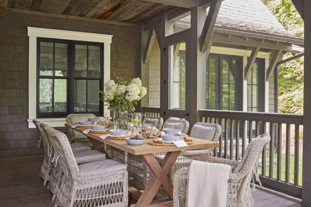 Covered porch with a wooden dining table set for eight, wicker chairs with striped cushions, and a large vase of white hydrangeas.