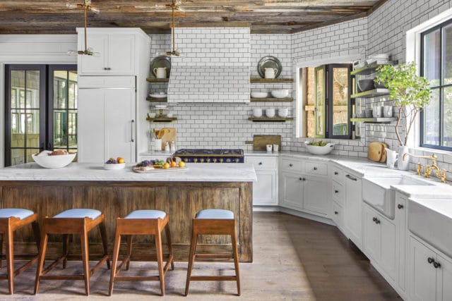 Rustic wood island with blue-cushioned stools in a white kitchen with subway tile backsplash, marble countertops, and a blue range.