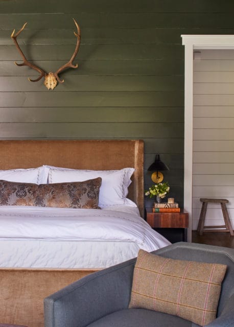 Bedroom with dark green shiplap walls, a king-sized bed with a brown velvet headboard, white bedding, and decorative pillows. A mounted deer head adorns the wall above the bed.