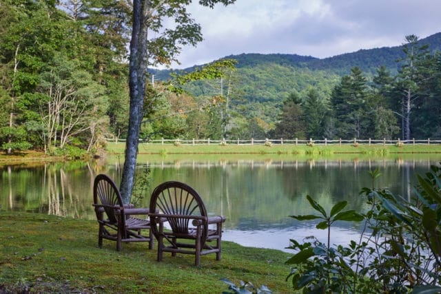 Two rustic wooden chairs sit on a grassy bank overlooking a calm lake with reflections of trees and a distant, tree-covered mountain under a cloudy sky.
