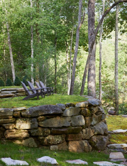Stone retaining wall with Adirondack chairs on a grassy knoll surrounded by trees.