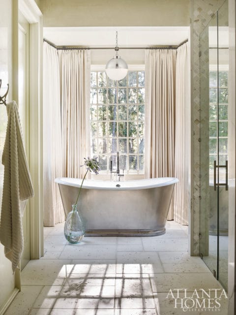 Freestanding bathtub in a bright bathroom with large window, sheer curtains, and tiled floor. A glass vase with a single purple flower sits on the floor.