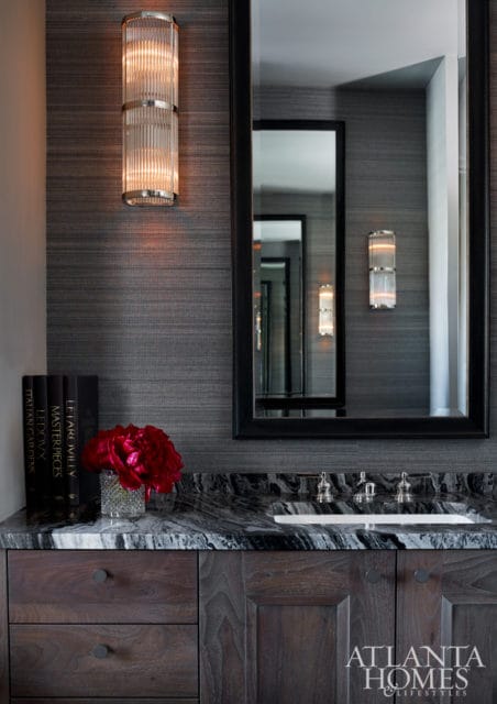 Dark wood vanity with marble countertop, glass vase with red flowers, and fluted glass wall sconces above a large black-framed mirror.