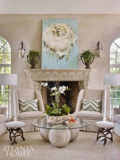 Living room with a fireplace, two upholstered chairs with green chevron pillows, a glass coffee table with a floral centerpiece, and a large abstract painting of a white flower above the mantel.