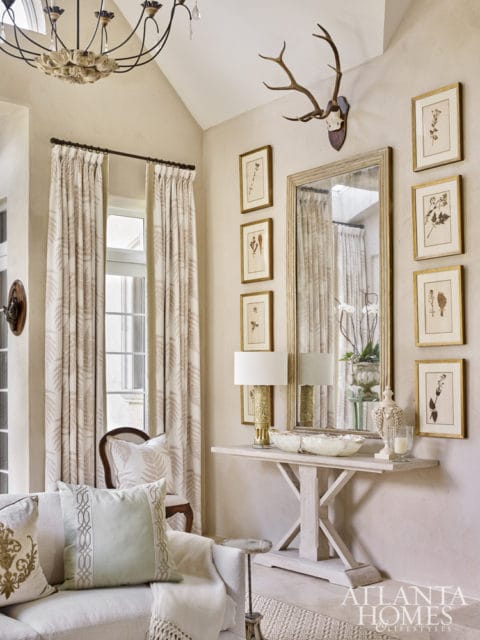 Living room with neutral-toned walls, featuring a large mirror, botanical prints, a console table with lamps and decorative bowls, and a sofa with decorative pillows.