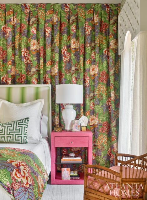 Bedroom with green floral curtains, a pink nightstand with a white lamp, and a bed with a green striped headboard and patterned bedding.