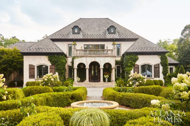 French-inspired estate with stucco facade, arched entryway, dormer windows, and manicured boxwood gardens with hydrangeas.