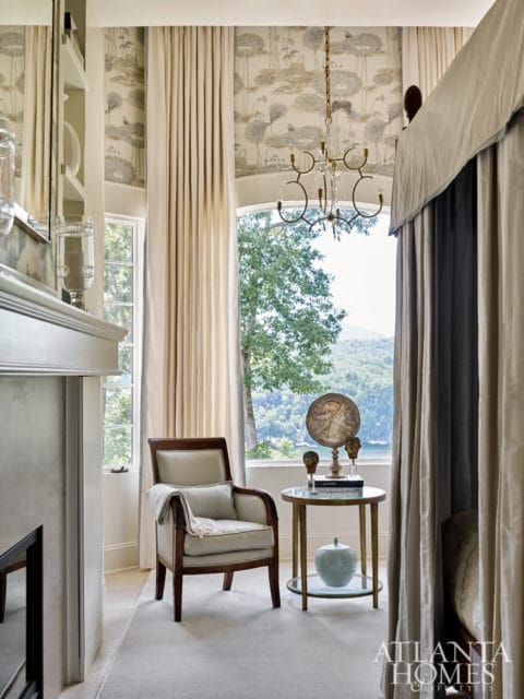 Bedroom with a four-poster bed, upholstered armchair, ornate chandelier, and scenic window views. The walls feature botanical wallpaper and floor-to-ceiling curtains.