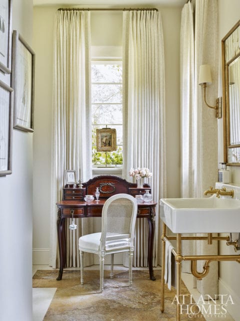 Powder room with a vintage wooden vanity, cane-backed chair, and a white porcelain sink with brass fixtures. Cream curtains frame a window overlooking greenery.