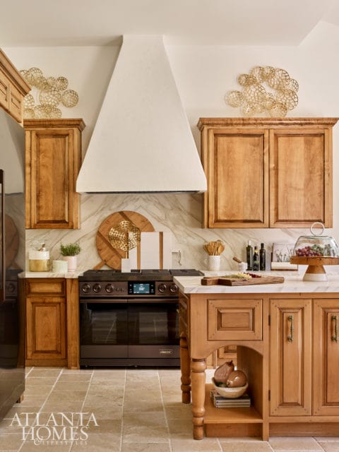 Warm wood cabinetry and a large white vent hood in a kitchen with marble backsplash and stone tile flooring. A dark range and a wooden island with a marble countertop are visible.