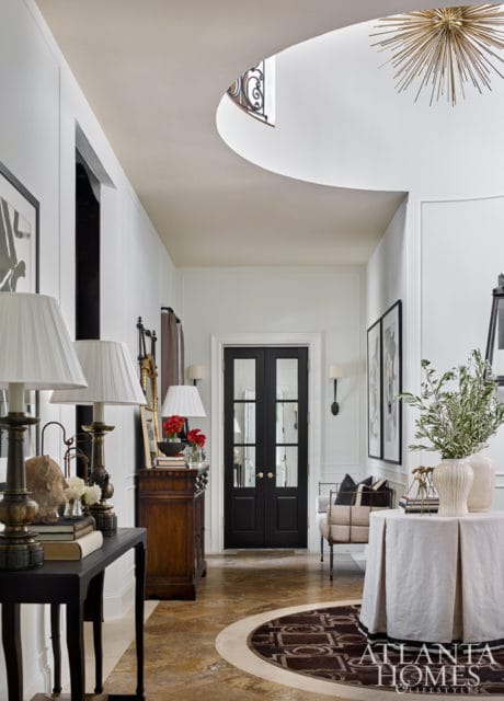 Hallway with white walls, black double doors, a curved ceiling with a skylight, and a starburst chandelier. Features include a dark wood console table with lamps, a round rug, and a draped table.