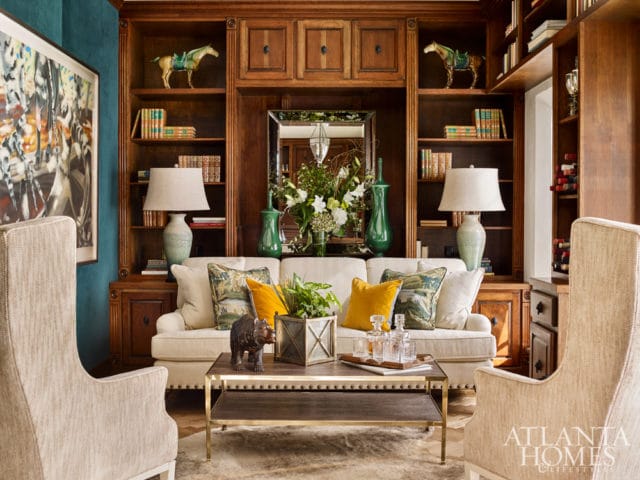 Living room with built-in wooden bookshelves, a cream sofa with decorative pillows, two upholstered armchairs, and a coffee table with decanters and a bear figurine.