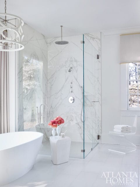 Modern white bathroom with a freestanding tub, marble-tiled walk-in shower with glass enclosure, and a clear acrylic chair with towels.