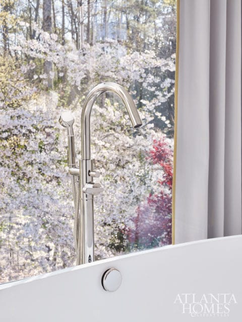 White bathtub with a chrome faucet and handheld showerhead, overlooking a window with blooming trees.