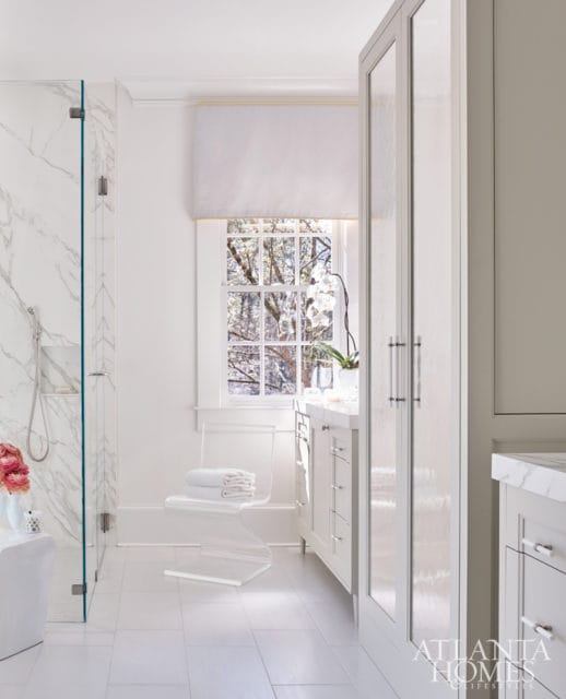 White bathroom with marble shower walls, a clear acrylic chair with stacked towels, and white cabinetry with marble countertops.