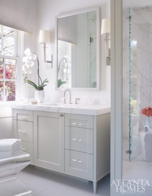 Light gray bathroom vanity with marble countertop, white orchid in a vase, and a large mirror above the sink. A glass shower door with blue accents is visible to the right.