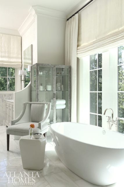 Light-filled bathroom with a freestanding white bathtub, a grey upholstered armchair, a glass-fronted cabinet filled with toiletries, and marble flooring.