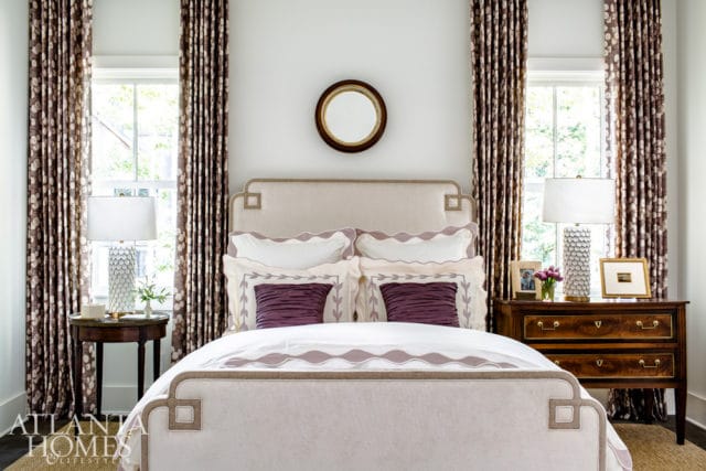 Bedroom with a cream upholstered headboard, purple accent pillows, and patterned drapes. A round mirror hangs above the bed.