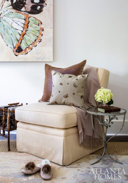 Cream upholstered ottoman with a brown velvet pillow and a decorative pillow featuring insects and butterflies, draped with a mauve throw blanket.