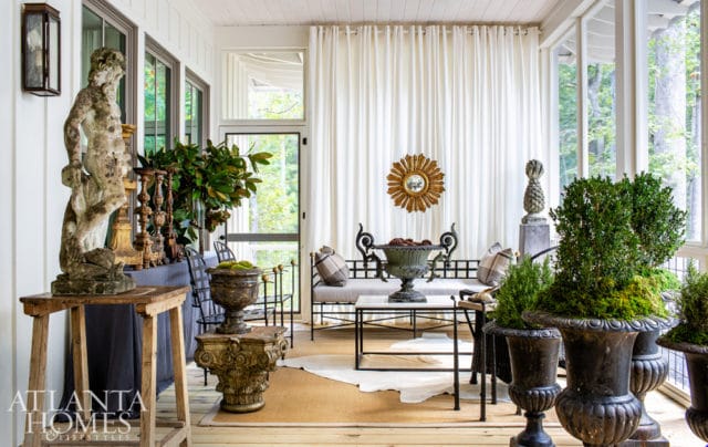 Screened porch with white curtains, wrought iron furniture, antique statues, and large planters filled with greenery.