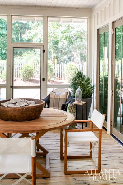 Screened porch with a round wooden dining table, director's chairs, and a wicker chair with a striped pillow. Large windows overlook a fenced yard with trees.