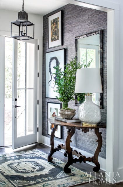 Entryway with textured gray wallpaper, a dark wood console table with a white ceramic lamp, a potted plant, and framed art. A patterned rug lies on the dark wood floor.