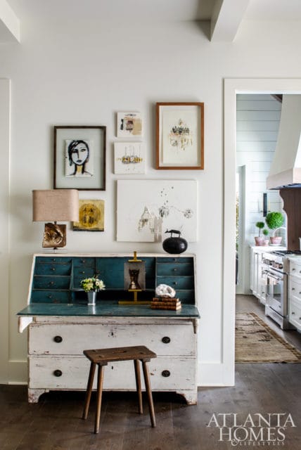 Secretary desk with distressed white and teal drawers, a wooden stool, and a collection of framed art on the wall above.