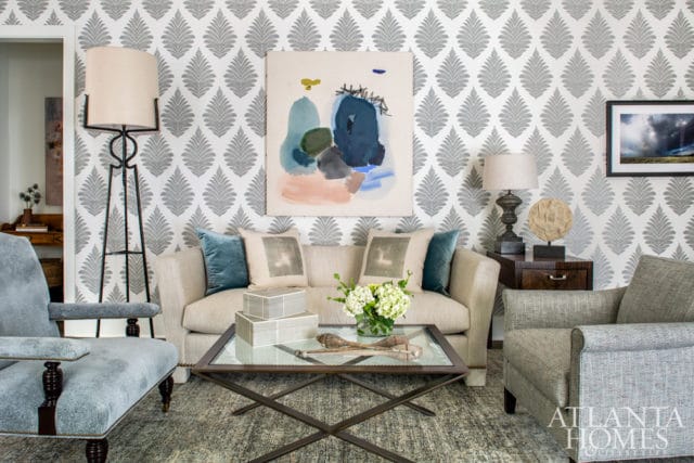 Living room with grey leaf-patterned wallpaper, a beige sofa with teal and grey pillows, a glass-top coffee table, and two armchairs.