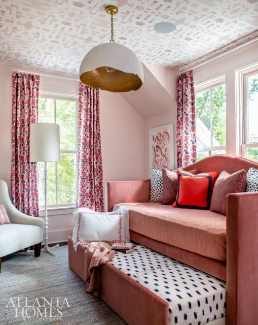 Pink daybed with a pull-out trundle bed in a room with patterned wallpaper, floral curtains, and a large pendant light.