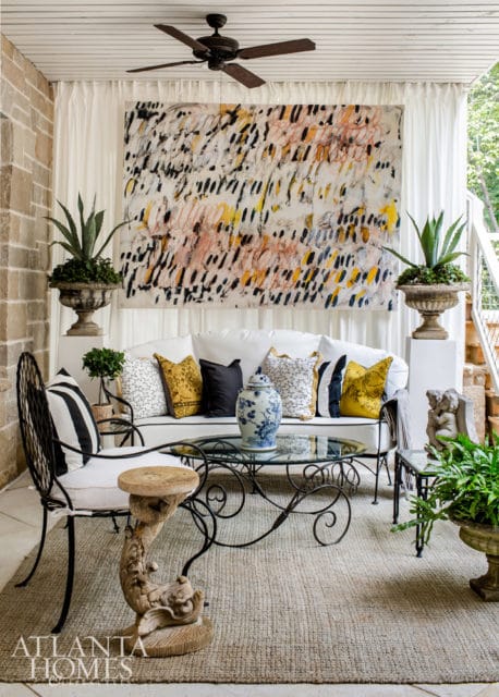 Covered patio with white sofa, black metal armchair, glass coffee table, and large abstract art. Stone walls and white curtains frame the space.