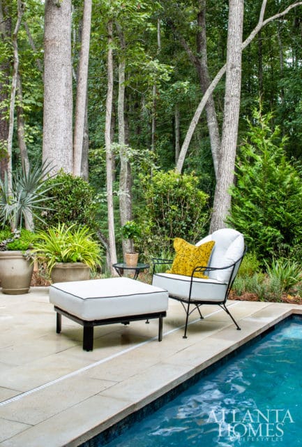 White outdoor seating with black trim and a yellow patterned pillow sits on a stone patio next to a pool, surrounded by lush greenery and trees.
