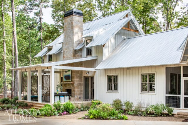 Modern farmhouse with white board and batten siding, metal roof, and stone chimney. Features a covered porch with decorative screens and landscaping.
