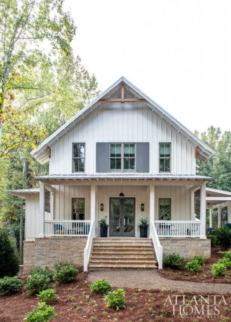 White farmhouse-style house with stone foundation, covered porch, and large gabled roof. Double glass doors lead to the entrance, flanked by windows and planters.