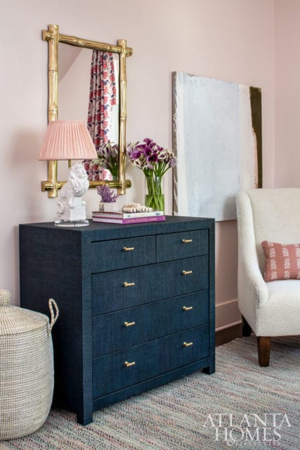 Dark blue dresser with brass hardware, pink pleated lamp, white lion sculpture, and a vase of purple flowers against a pale pink wall.