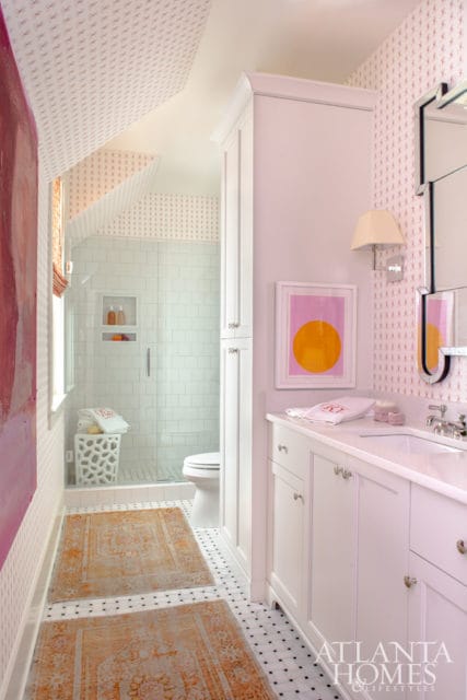 Pink and white patterned wallpaper adorns this bathroom with a white vanity, glass-enclosed shower with white subway tile, and two orange patterned rugs on a black and white tiled floor.