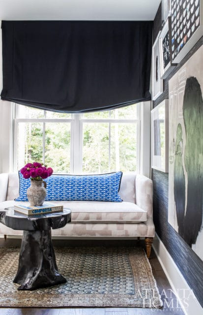 Living room with a light-colored sofa featuring a blue patterned lumbar pillow, a dark wood stump side table with a vase of purple flowers and a book, and artwork on a dark textured wall.