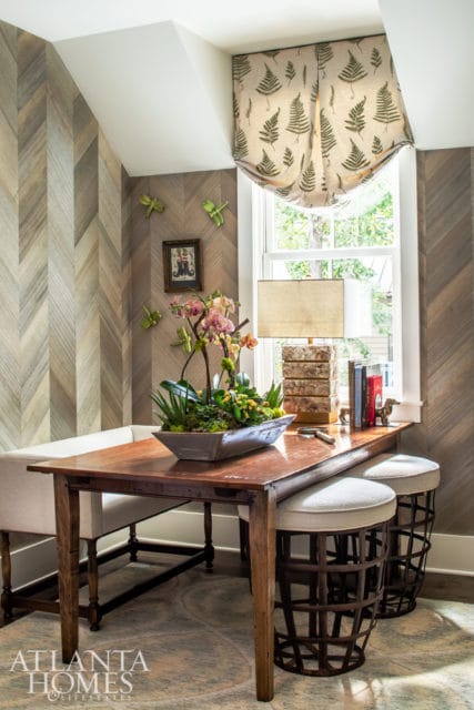 Wooden desk with a plant centerpiece, orchid arrangement, lamp, and books. Walls feature chevron wood paneling and a fern-patterned window shade.