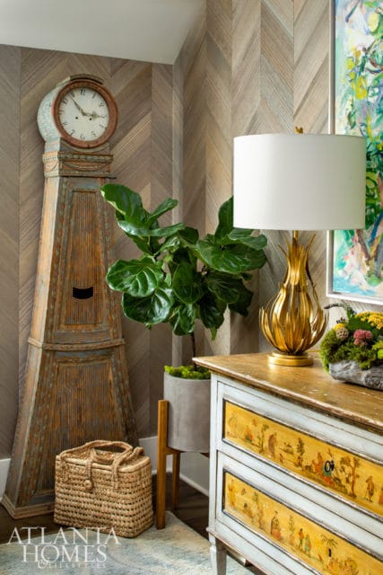 Corner of a room with a tall antique clock, a fiddle leaf fig plant in a concrete planter, and a chest of drawers with hand-painted scenes.