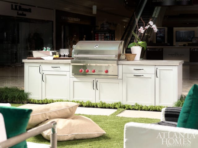 White outdoor kitchen island with a stainless steel grill, concrete countertop, and storage cabinets, set up on artificial grass with pillows and plants.