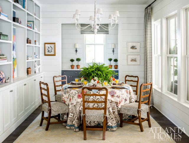 Dining room with a round table covered in a floral and bird print tablecloth, surrounded by wooden chairs with patterned cushions. White built-in shelving lines one wall.