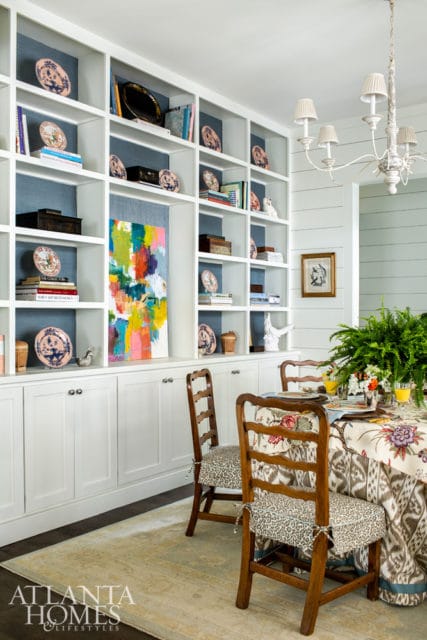 Dining room with built-in white bookshelves filled with decorative plates and books, a colorful abstract painting, and a dining table set with a floral tablecloth.